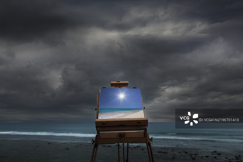 在阴天的海滩上，画架上展示着阳光下的热带海滩画作。图片素材