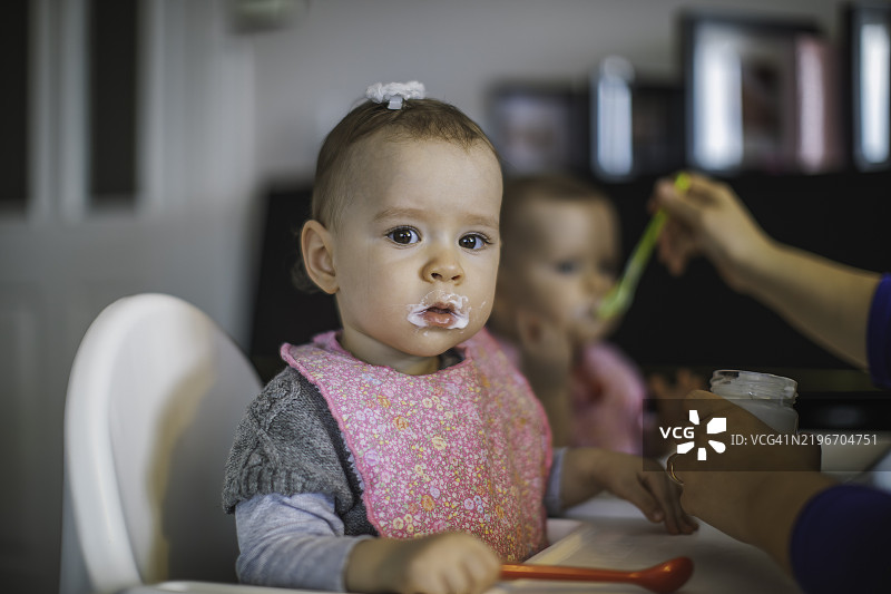 母亲在家中给双胞胎宝宝喂食，宝宝一边吃一边看着镜头图片素材