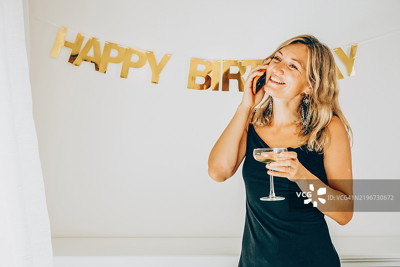 穿着黑色连衣裙的快乐美丽女性，喝着香槟，打电话并听取祝贺。图片素材