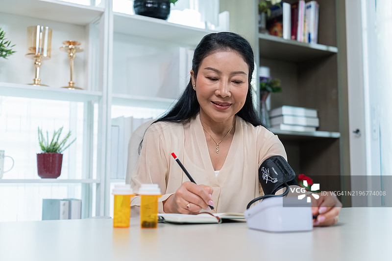 一位穿着浅米色 blouse 的老年女性微笑着，轻柔地在笔记本上记录她的读数，突显了她对健康生活方式和福祉的承诺，强调了日常生活中定期检查和自我护理的重要性。图片素材