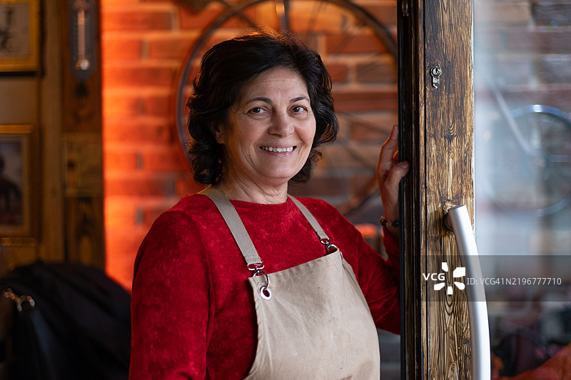 快乐的成熟的女性企业家穿着围裙，微笑着欢迎顾客来到她的小店。图片素材