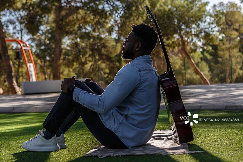 一个背光的男人坐在公园里，手里拿着一把木吉他，周围是树木。图片素材