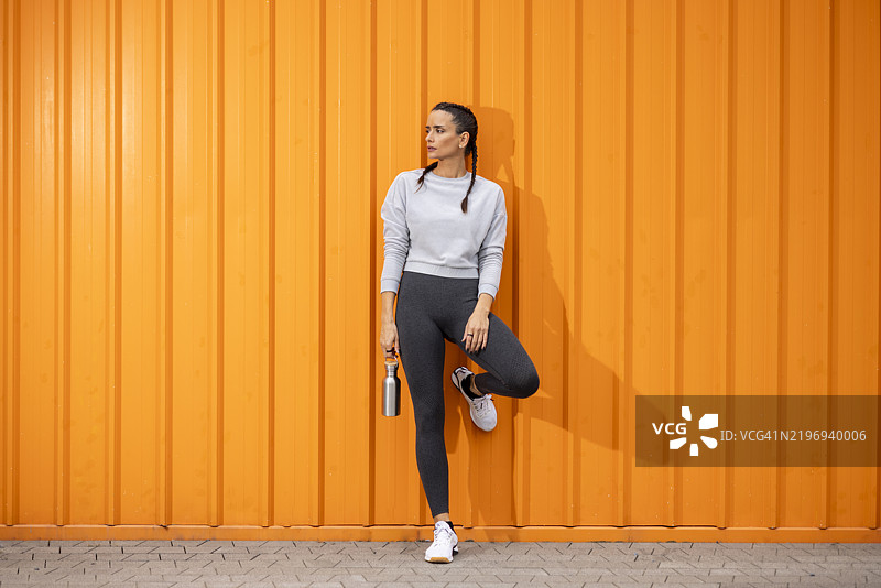 运动女性手持可重复使用的水瓶，摆姿势于橙色墙壁前图片素材
