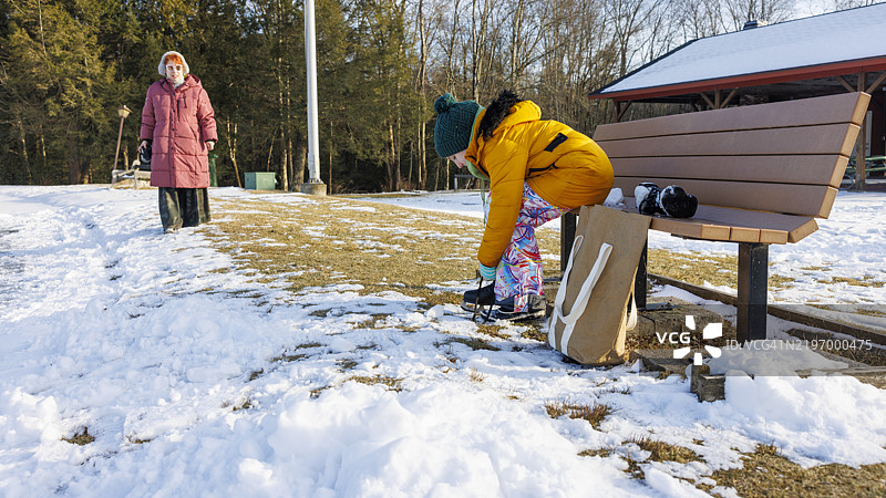 母女在冰封湖面上准备滑冰。女孩系着冰鞋，而红发妈妈在一旁观看。图片素材