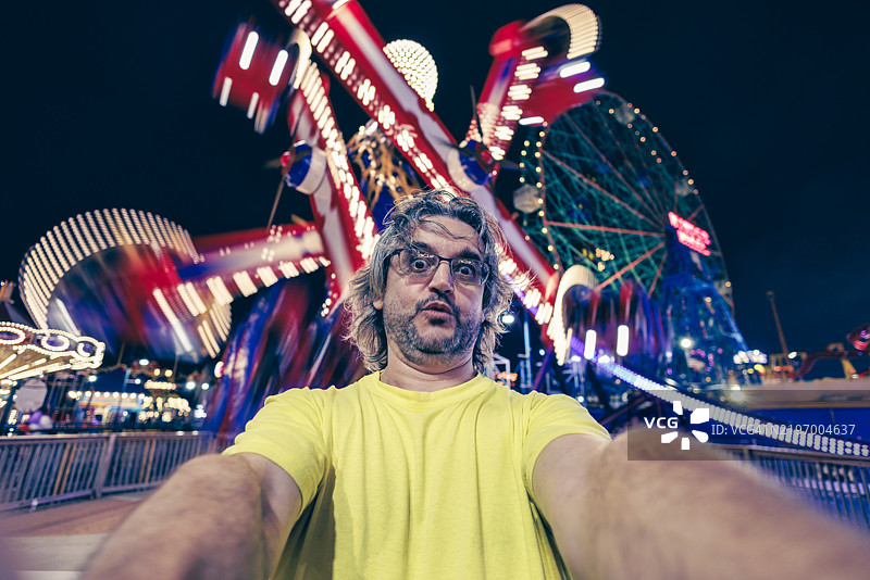 惊讶的成熟游客戴着眼镜，在夜间游乐园拍自拍照图片素材
