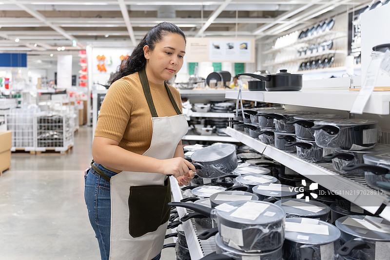 一位中年亚洲女性，作为小企业主，穿着围裙正在整理商店货架上的炊具库存。图片素材