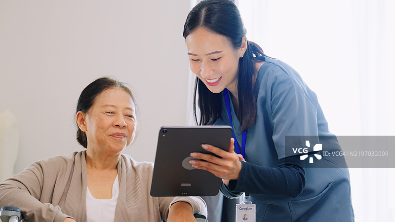 亚洲护理人员穿着蓝色护士服，向退休的老年女性解释医疗程序和医疗记录，使用数字平板电脑，展示了技术在病人护理中的整合以及在医疗环境中清晰沟通的重要性。图片素材