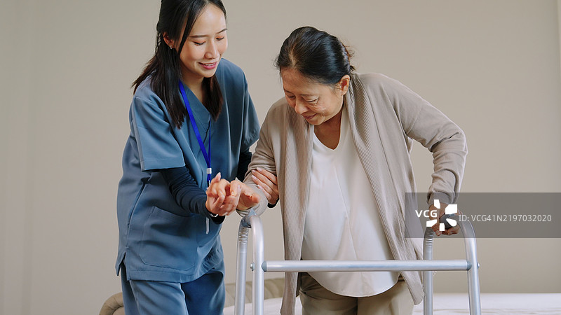 穿着蓝色护士服的亚洲女性护理人员正在帮助一位老年女性使用助行器，协助她从助行器站起，过渡到床上休息，支持她的健康和生活方式。图片素材