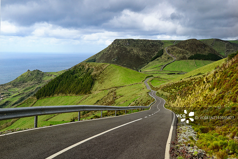 一条蜿蜒的道路穿过郁郁葱葱的山丘，远处的海洋在阴云密布的天空下展现出迷人的景色。图片素材