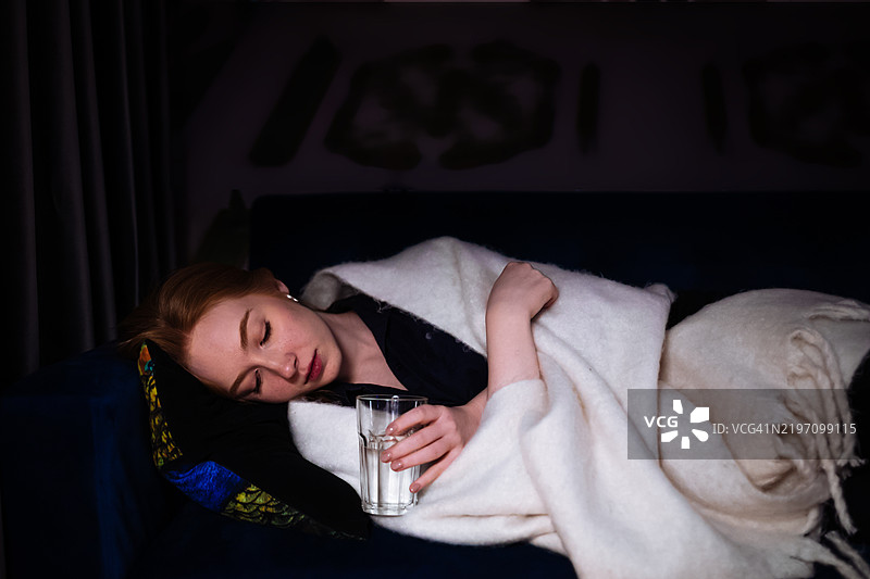 一位女性靠在沙发上，盖着毯子，手中握着一杯水，流露出疲惫与宁静的感觉。柔和的灯光增添了平静的氛围。图片素材