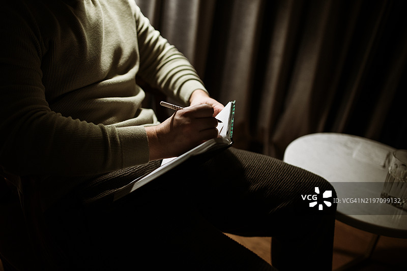 一位心理学家在笔记本上写字，画面传达出专注和在柔和灯光下进行会谈时细致记录的重要性。图片素材