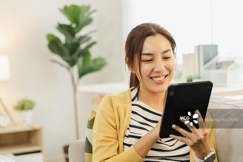 微笑的女性在舒适的沙发上放松，享受使用平板电脑的乐趣。非常适合生活方式和科技内容。图片素材