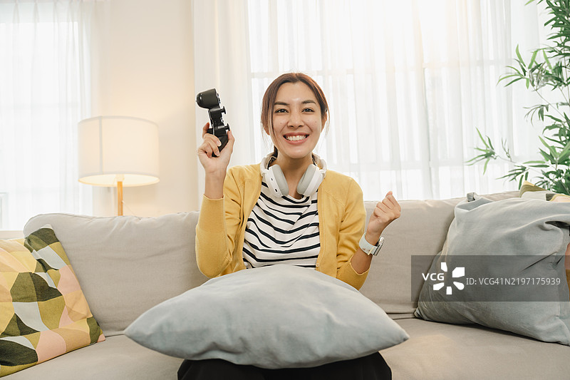 年轻女性兴奋地握着游戏手柄，坐在舒适的沙发上，耳机挂在脖子上，享受在家玩游戏的乐趣。图片素材