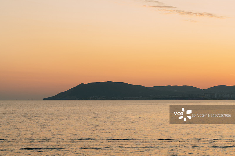 温暖的日落色彩映照在平静的海面上，山影若隐若现。图片素材