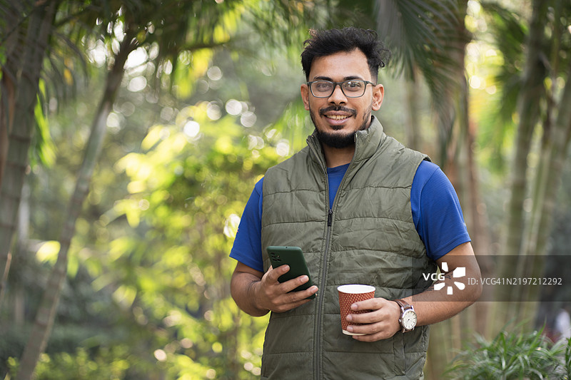 自然中悠闲的年轻男子，手持咖啡和智能手机图片素材