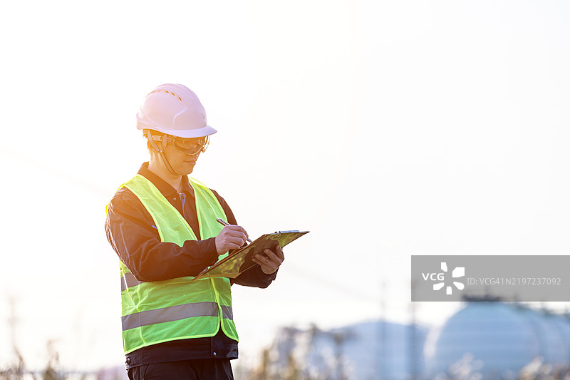男性首席工程师穿着安全背心、硬帽和护目镜，在现代联合循环发电厂进行测量记录。图片素材