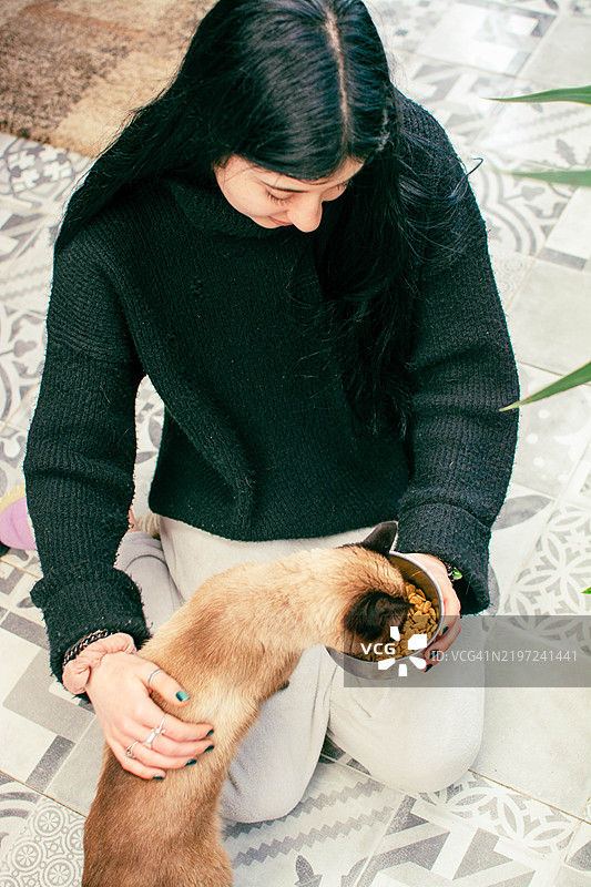 年轻女性在家里喂她的暹罗猫图片素材