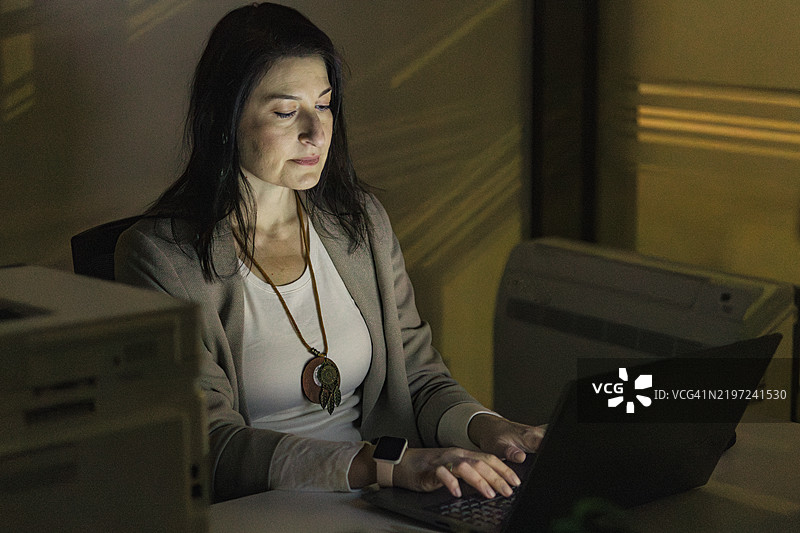 疲惫的中年女商人在办公室加班。她独自一人，使用笔记本电脑，因夜间工作而感到疲惫。图片素材
