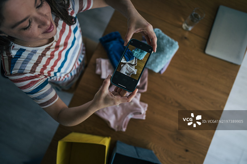 母亲坐在桌子旁，专注于用手机拍摄衣物。她小心翼翼地调整相机角度，捕捉面料或设计的细节。图片素材