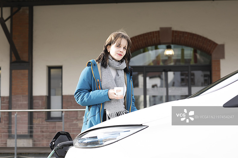 冬季城市环境中，穿着蓝色外套的女性手持杯子，站在一辆电动车前，展现环保理念。图片素材