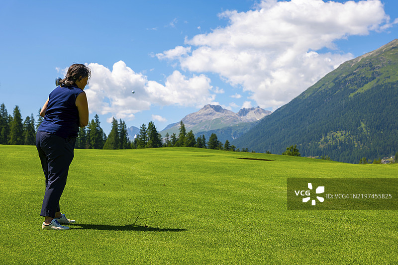 在瑞士格劳宾登州的马洛伊亚、圣莫里茨的恩加丁，阳光明媚的夏日，蓝天白云下，一位女性正在打高尔夫球，背景是壮丽的山景。图片素材