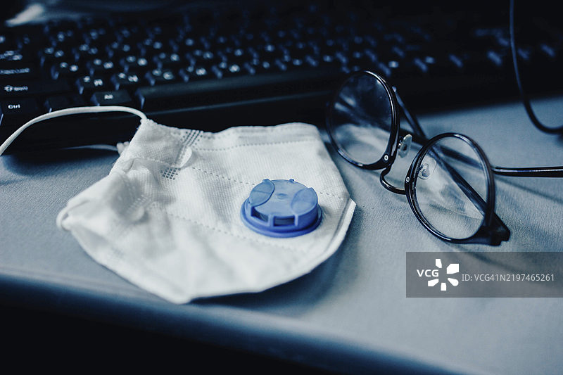 一只带蓝色阀门的白色口罩和眼镜放在键盘旁的桌子上图片素材