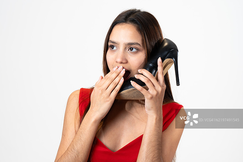 惊讶的年轻女性在白色背景下用高跟鞋当电话图片素材