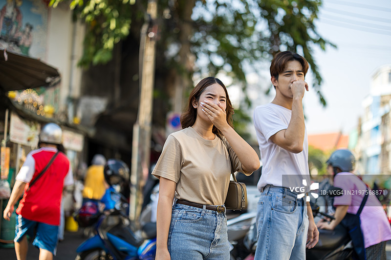 两位亚洲旅行者在拥挤的城市中因严重的空气污染而捂住鼻子，周围有浓厚的雾霾和交通。图片素材