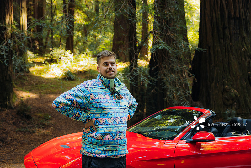 快乐的男人站在红色敞篷车旁，身处红木森林中图片素材