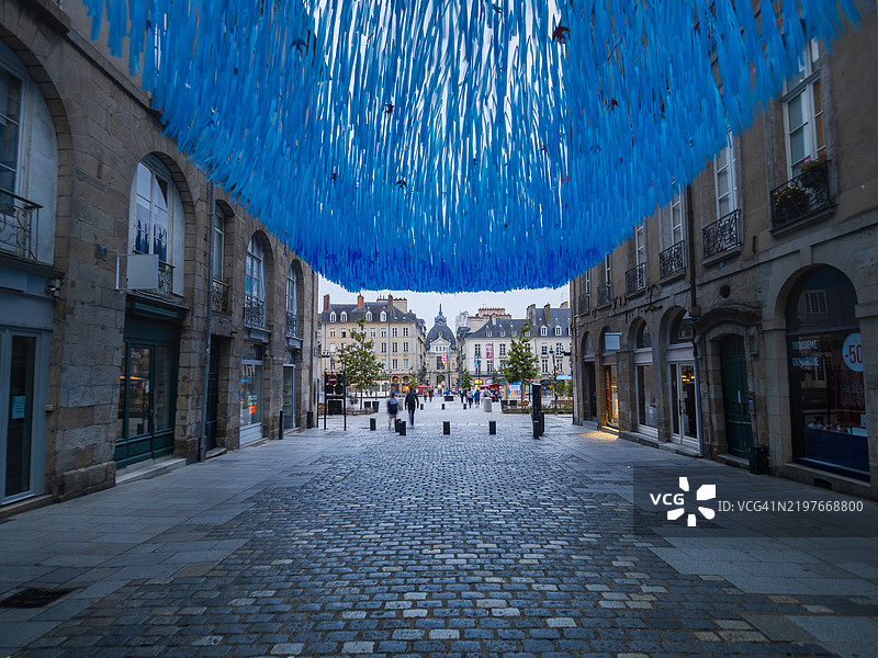 雷恩历史区的一条鹅卵石步行购物街的广角视图,街道两旁是商店和零售店,覆盖着蓝色丝带,尽头是市政厅广场,位于伊勒-维莱讷省 - 布列塔尼(法国)。图片素材