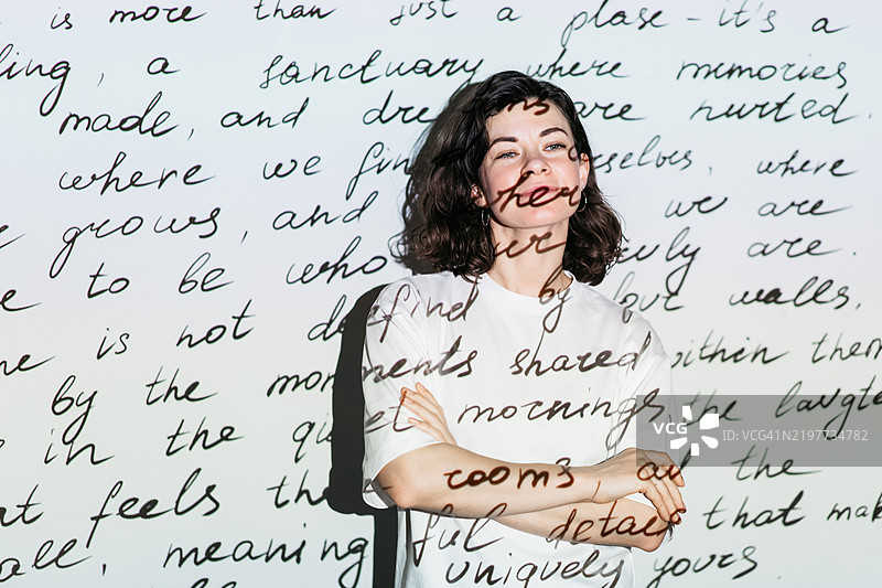 一位女性作家站在白色墙壁前，艺术作品的投影映射在她和墙上。图片素材