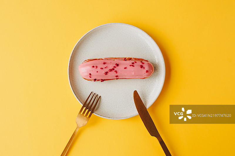 极简主义的餐桌布置，白色盘子中央放着一块粉色糖霜的泡芙，旁边是金色的刀叉，背景为明亮的黄色。图片素材