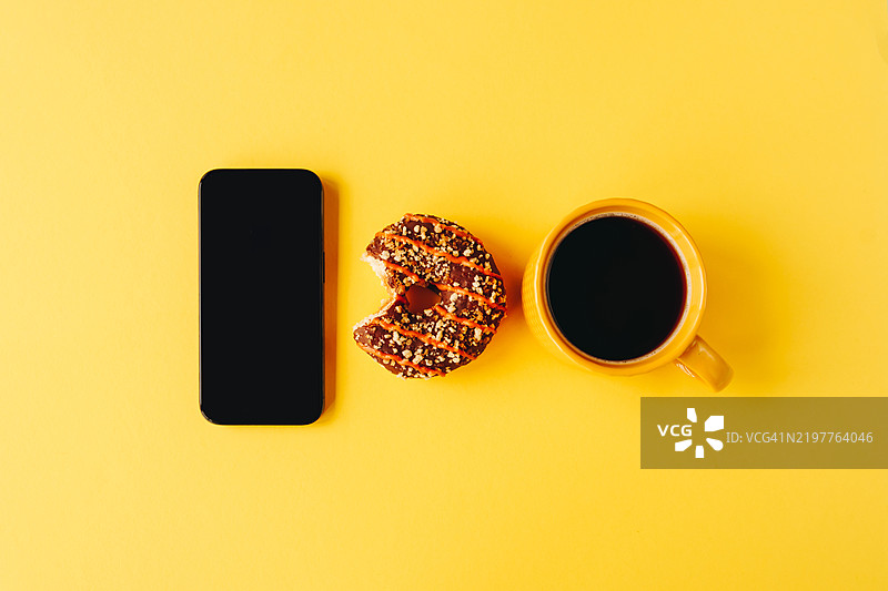 一幅生动的作品，展示了一只黄色咖啡杯、一块被咬了一口的巧克力淋酱甜甜圈，以及一部时尚的黑色智能手机，背景为明亮的黄色。图片素材