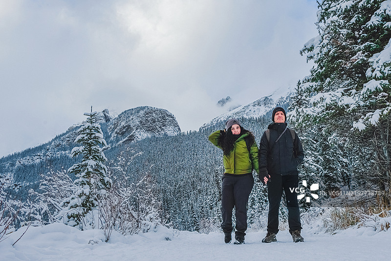一对恋人手牵手，面向镜头，站在湖边，四周被山脉和雪覆盖，晴空万里，位于加拿大班夫。图片素材