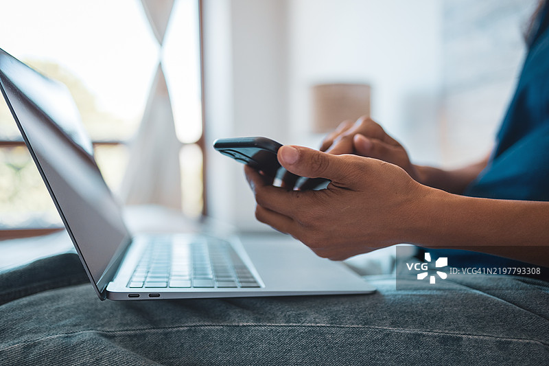在家环境中，一个人正在使用智能手机和笔记本电脑进行多任务处理的特写镜头图片素材