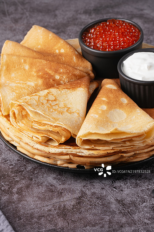 新鲜制作的煎饼高高叠起，上面放着红鱼子酱，旁边有碗酸奶油。图片素材