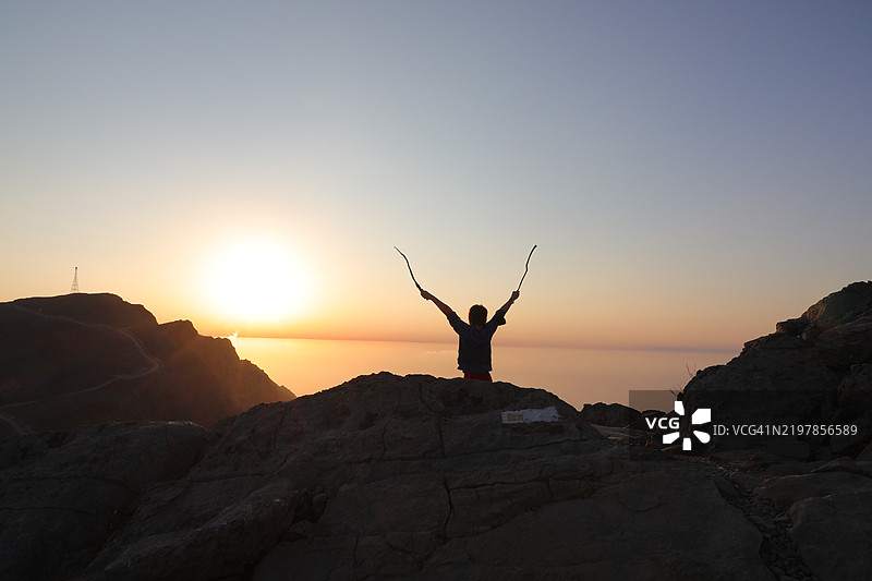 在日落时分，站在岩石上，双臂高举的男人剪影与天空相映成趣图片素材