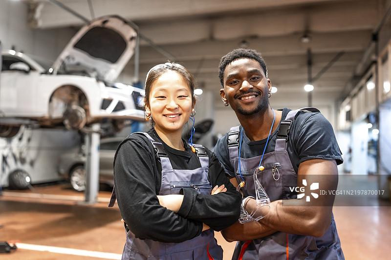 两位微笑的机械师交叉着手臂，展现出自信和专业，在一个充满工具和设备的繁忙汽车服务站内。图片素材