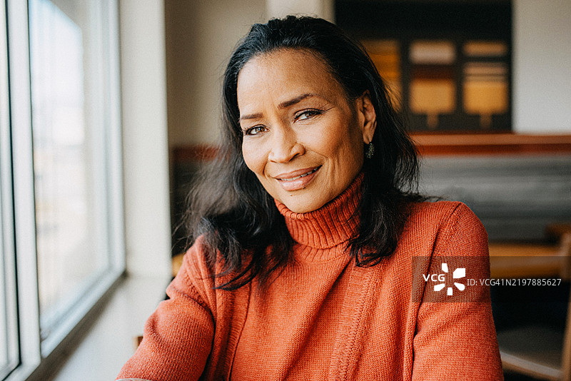 一位穿着舒适红色高领毛衣的女性坐在窗边，微笑着看向镜头。图片素材
