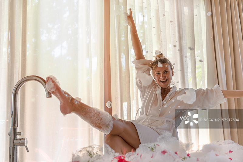 年轻美丽的女性穿着白色浴袍，坐在浴缸里向空中抛洒浴泡，现代浴室内有大窗户和米色窗帘。图片素材