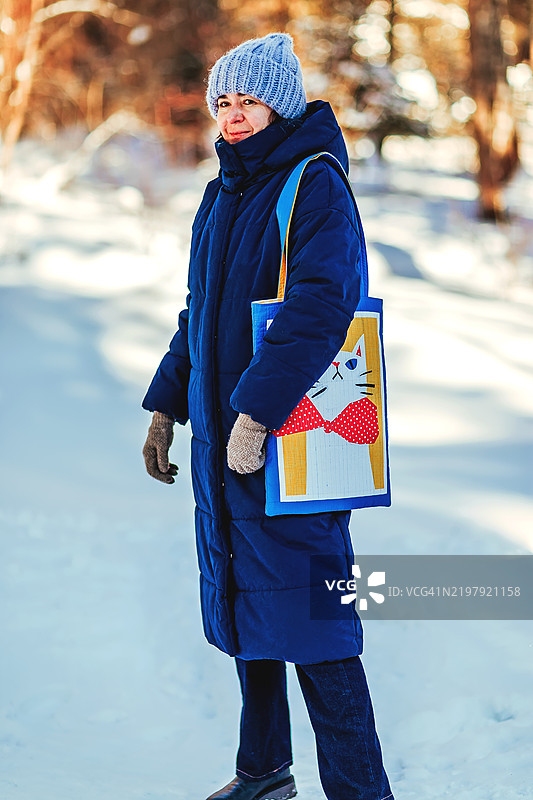 穿着冬季服装的女性在雪地中提着猫图案的拼布手提袋。一位穿着蓝色冬季外套、针织帽和手套的女性走在雪景中，手提一款时尚的猫主题拼布手提袋，温暖与时尚兼具。图片素材