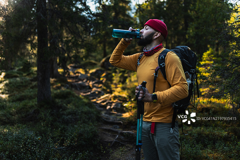 男子在森林中徒步旅行时，穿着冬季装备，背着背包，喝水。图片素材