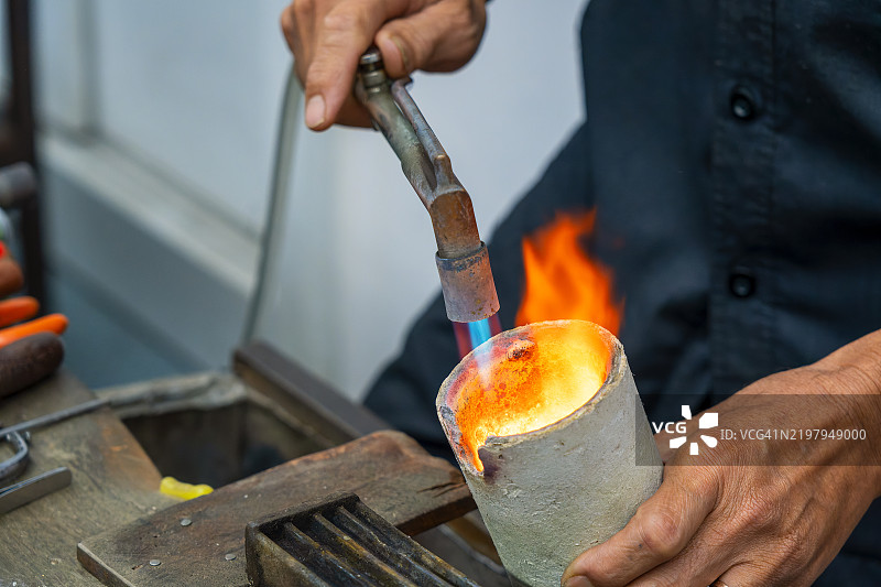 泰国曼谷的传统工作室中，银匠的手在用喷灯熔化银料，为制作珠宝做准备，展示了珠宝制作的复杂过程。图片素材