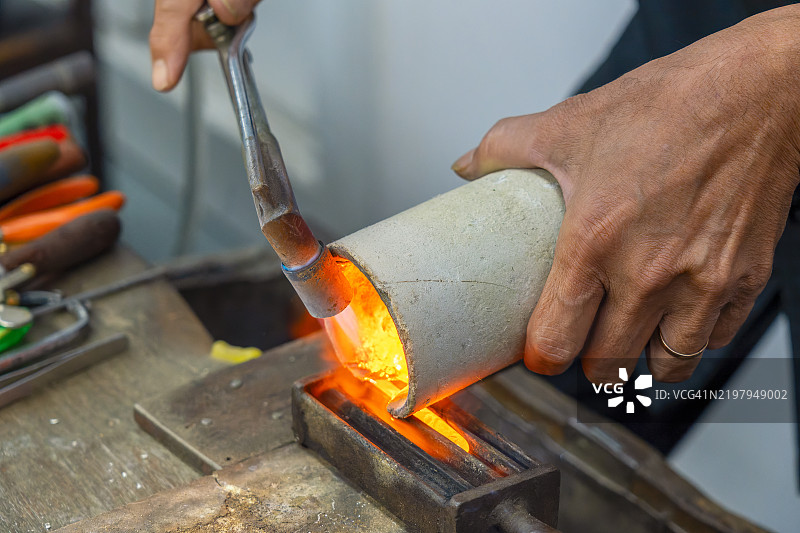 珠宝匠的手特写，使用火焰喷 torch 融化黄金，为在泰国传统工作室制作珠宝做准备，然后将黄金倒入模具中制作手工黄金珠宝。曼谷，泰国图片素材