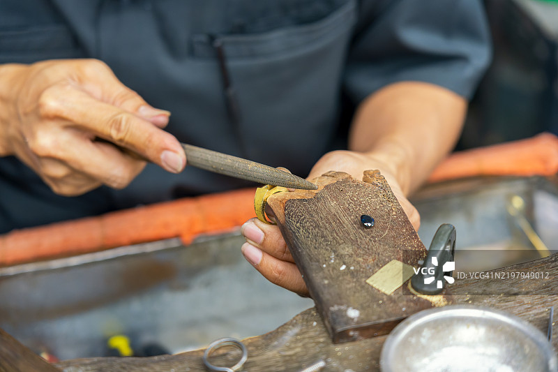 金匠的手特写，使用锉刀打磨、抛光和精细处理一枚金色银戒，为镶嵌宝石做好准备，背景为传统工坊。泰国曼谷图片素材