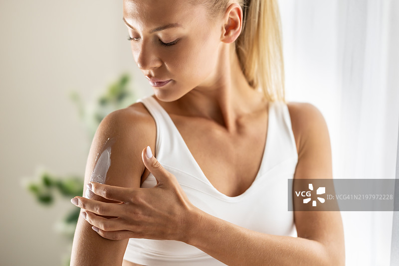 美丽的年轻女性在卧室里给肩膀涂抹护肤霜。护肤、水疗、健康和化妆品概念。图片素材