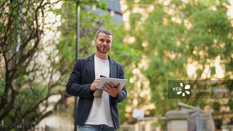 自信的年轻职业商人在城市环境中手持数字平板和可重复使用的水瓶的肖像图片素材
