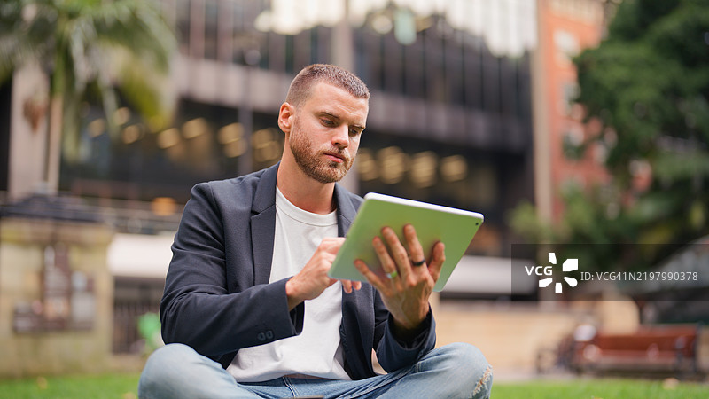 年轻的职业商人在城市公园的草地上坐着，使用数字平板电脑图片素材