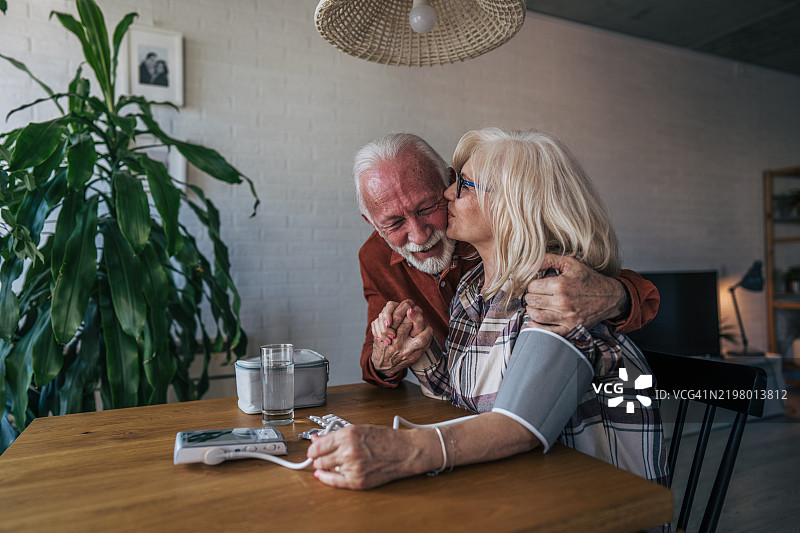 一对老年夫妇坐在桌子旁，女人正在测量她的血压。图片素材