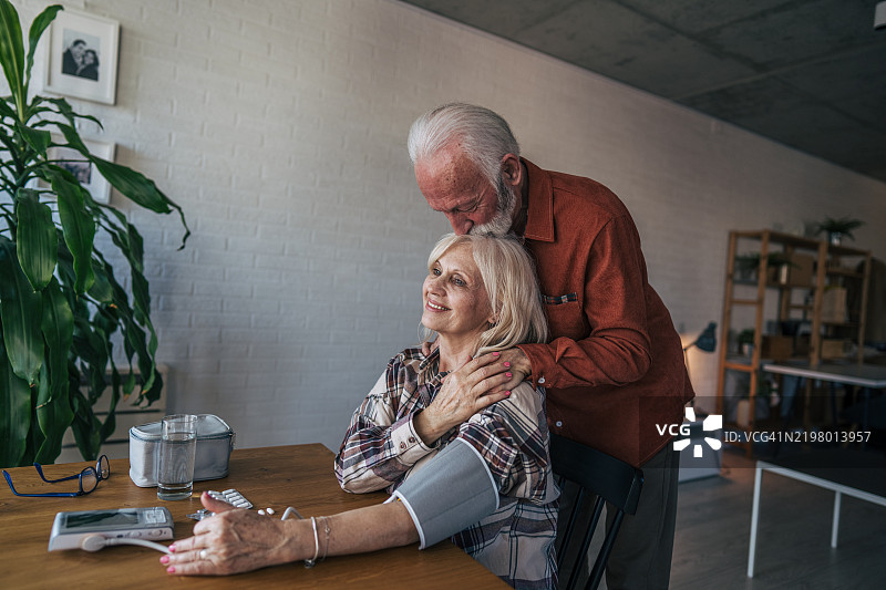 一位年长女性坐在桌子旁，用数字血压计测量自己的血压。她的丈夫站在她身后，轻轻吻着她的头。图片素材
